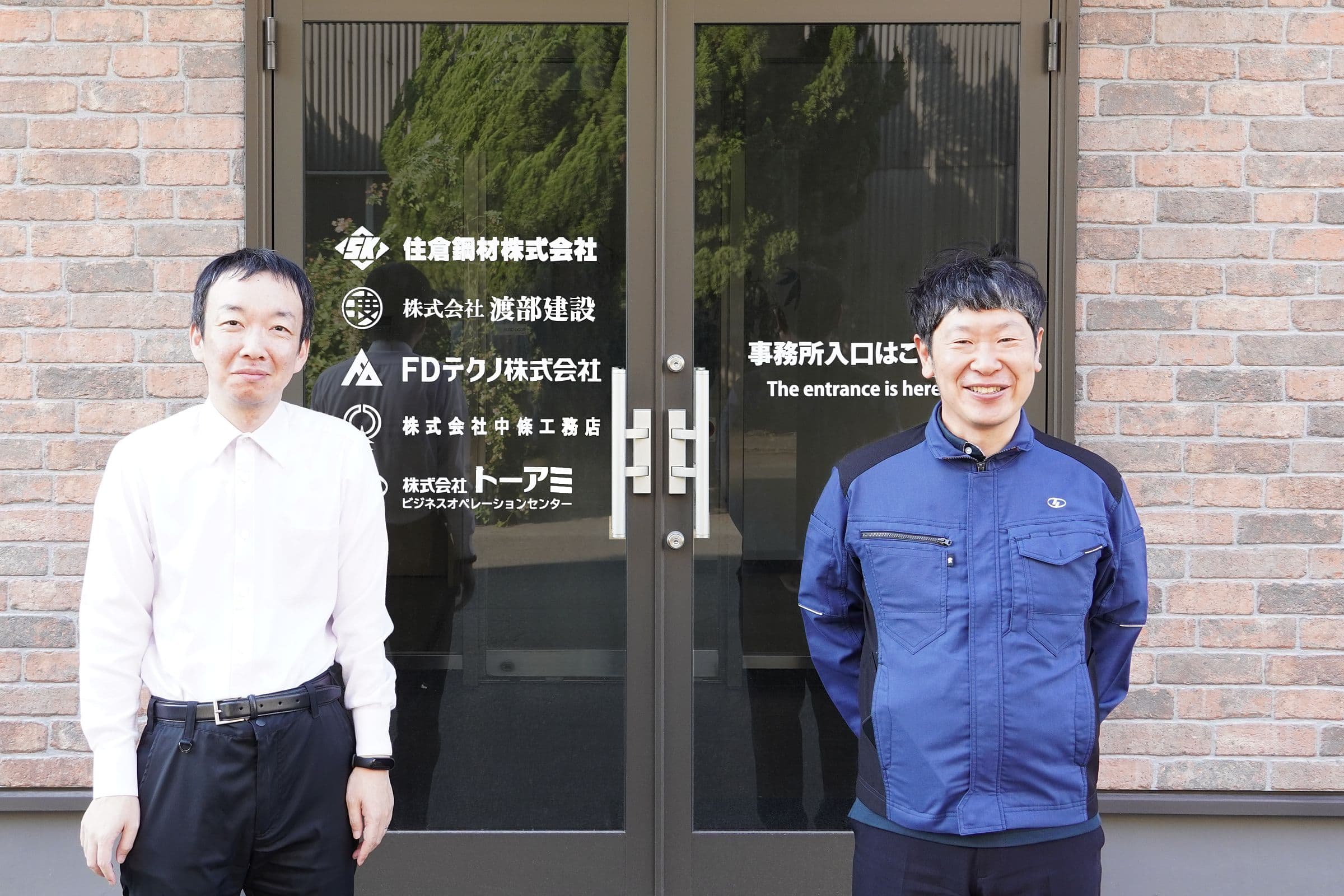 グループ各社の社名が入った北九州市小倉北区の拠点の入口に立つ伊藤和俊氏（右）